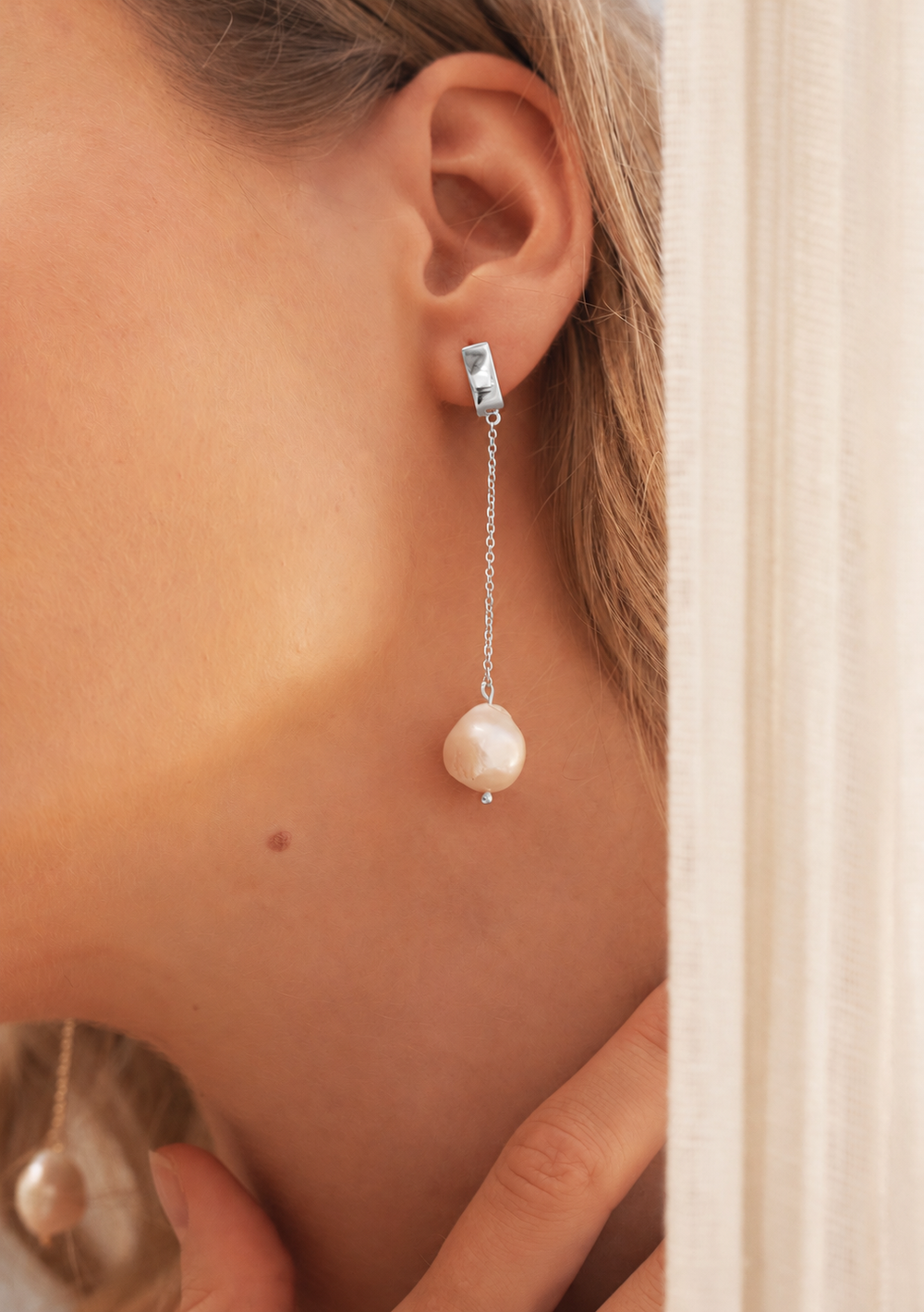 A close-up of a model's ear wearing a silver drop earring with a barbell-shaped stud and a dangling baroque pearl.