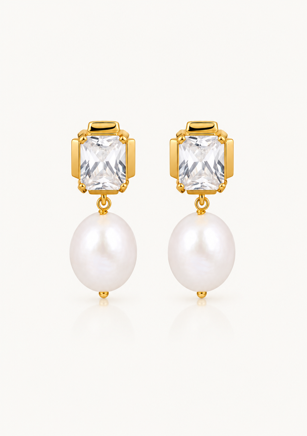 A pair of gold drop earrings featuring a rectangular clear faceted stone and a dangling white pearl, shown against a light off-white background.