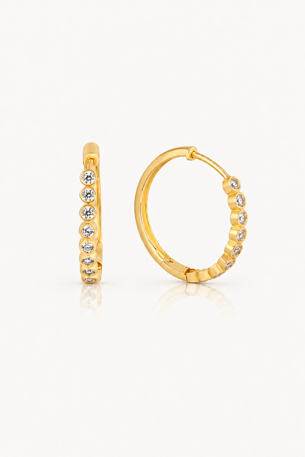 A pair of gold huggie hoop earrings with small clear round-cut stones set along the front, shown on a clean white background with a soft reflection.