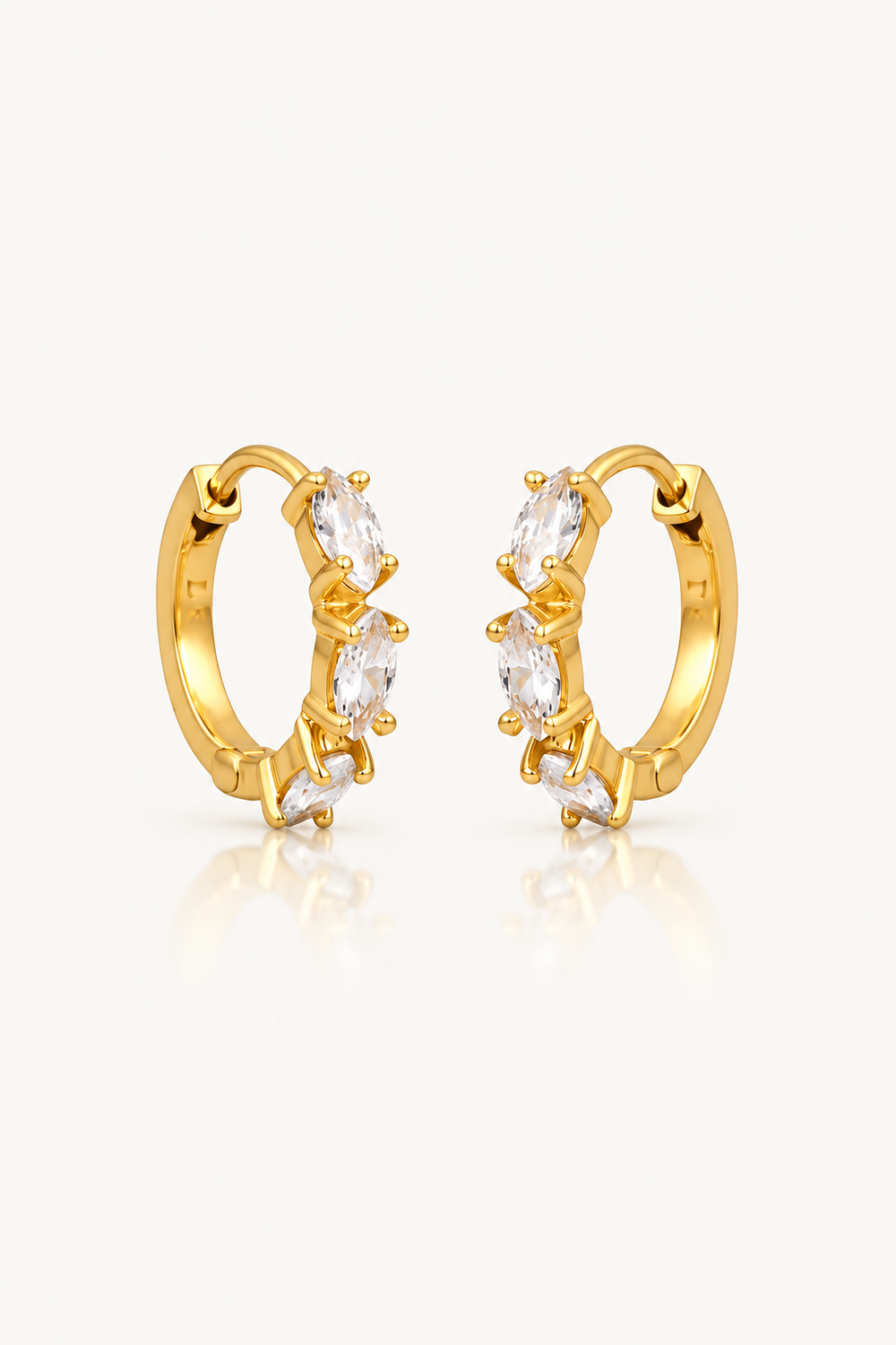 A pair of gold huggie hoop earrings with marquise-cut clear crystals, displayed on a clean off-white background with a soft reflection below.