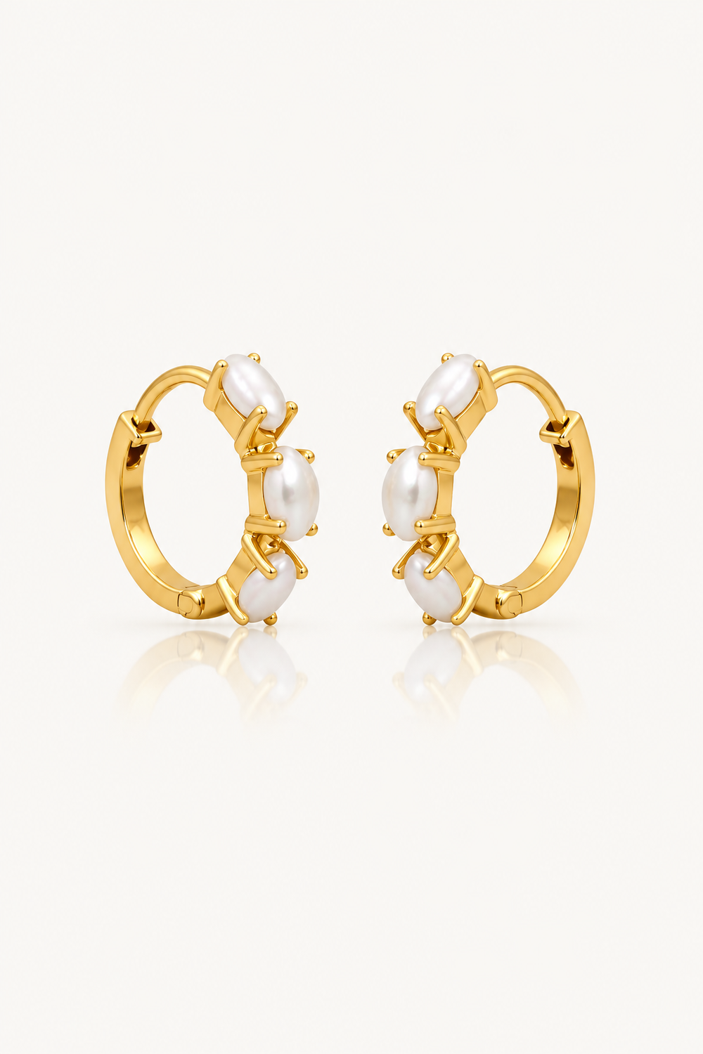 A pair of gold huggie hoop earrings with white faux-pearl bead details, displayed on a clean off-white background with a soft reflection below.