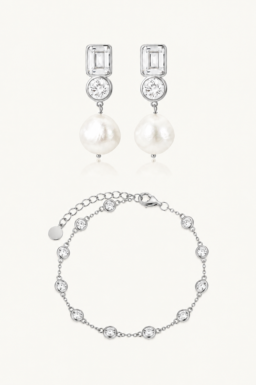 A modern jewelry bridal jewellery set featuring a pair of silver drop earrings with rectangular clear stones, a round clear stone, and a white pearl, paired with a matching silver chain bracelet adorned with clear round stones, on a light cream background.