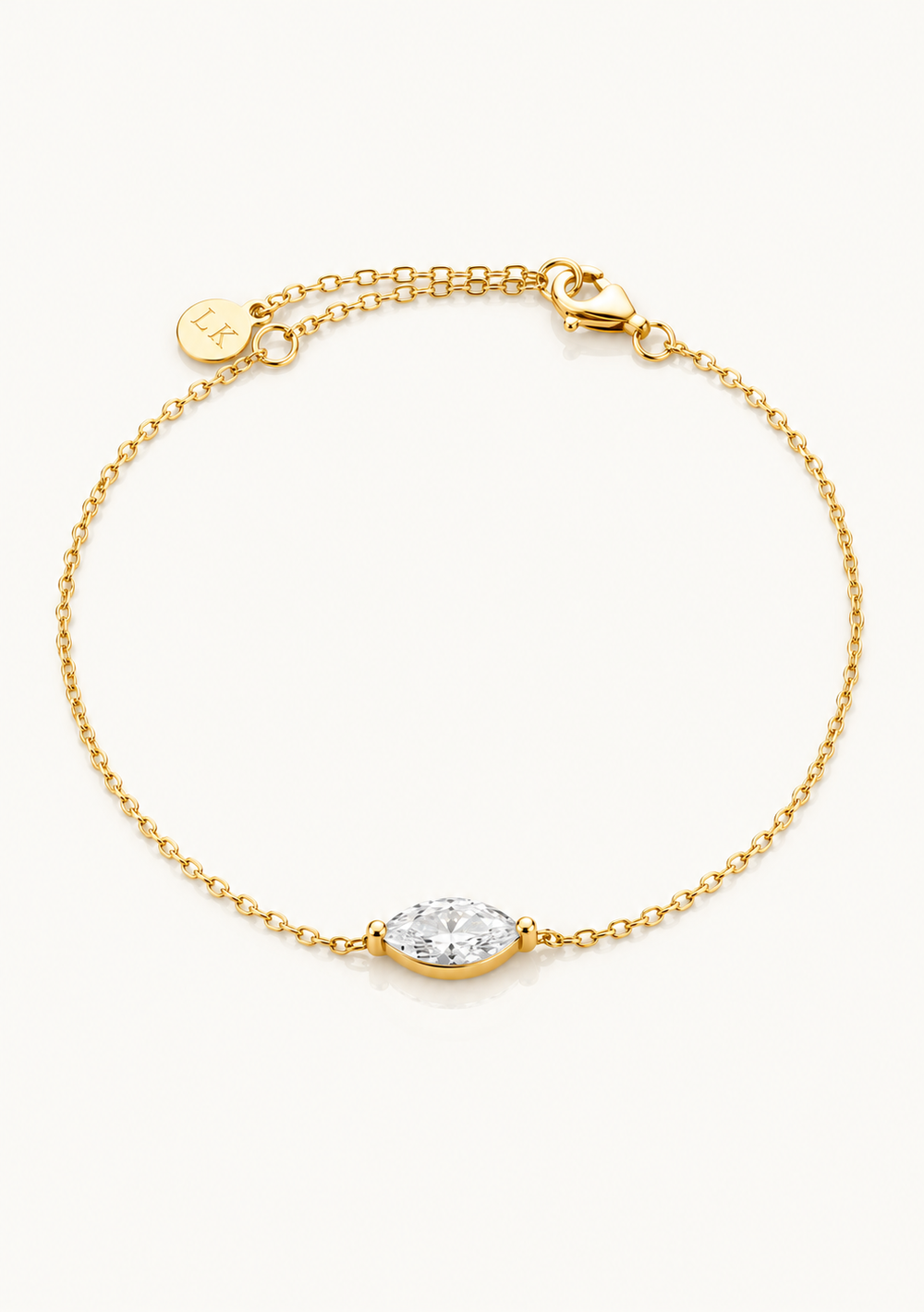 A delicate gold chain bracelet featuring a marquise-cut clear gemstone in a gold bezel setting, displayed on a clean off-white background. The bracelet includes an adjustable lobster clasp, a heart-shaped charm, and a circular brand tag engraved with 'L.