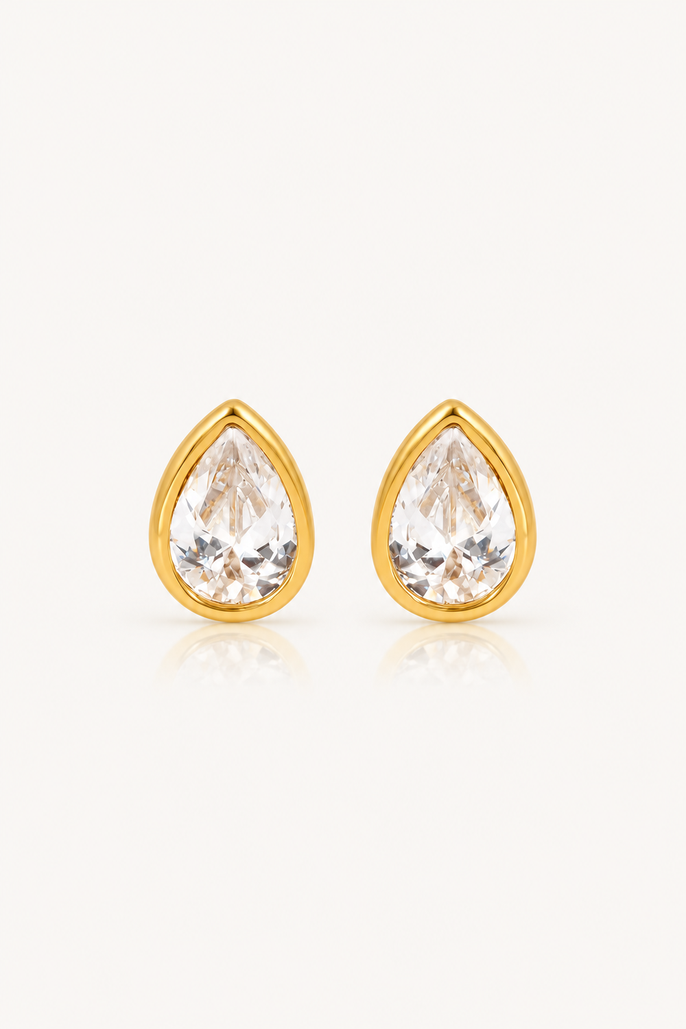 A pair of gold teardrop stud earrings with clear faceted gemstones, set against a plain off-white background with a soft reflection below.