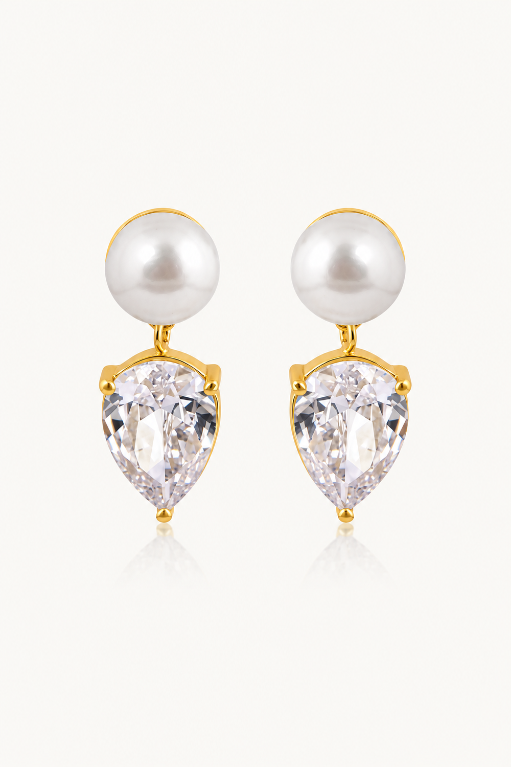 A pair of gold pearl drop wedding earrings featuring a white pearl and a clear pear-cut crystal, set against a light off-white background.
