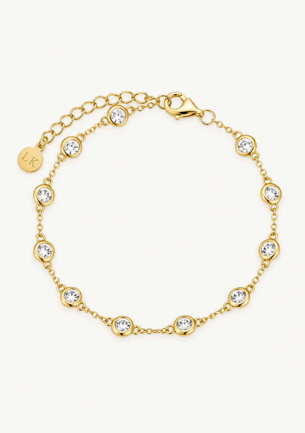 A gold chain bracelet with multiple bezel-set clear round crystals and a small round engraved charm, displayed on a light cream background.