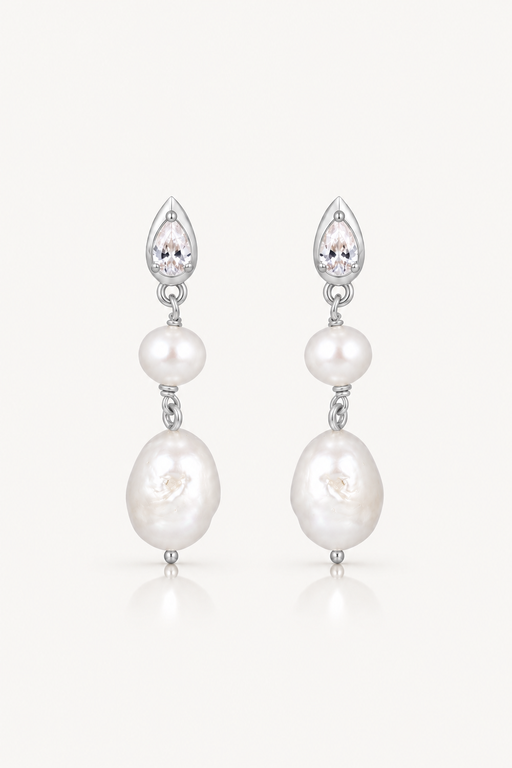 A pair of silver drop earrings featuring a teardrop-shaped clear crystal, a small white pearl, and a larger irregular white baroque pearl, set against a light off-white background.