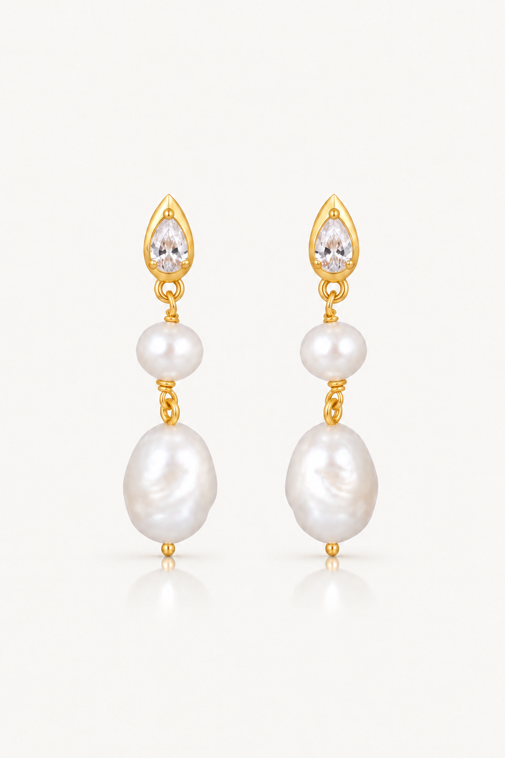 A pair of gold drop earrings featuring a small clear teardrop crystal, a white pearl bead, and a larger irregular white baroque pearl, shown on a light off-white background.