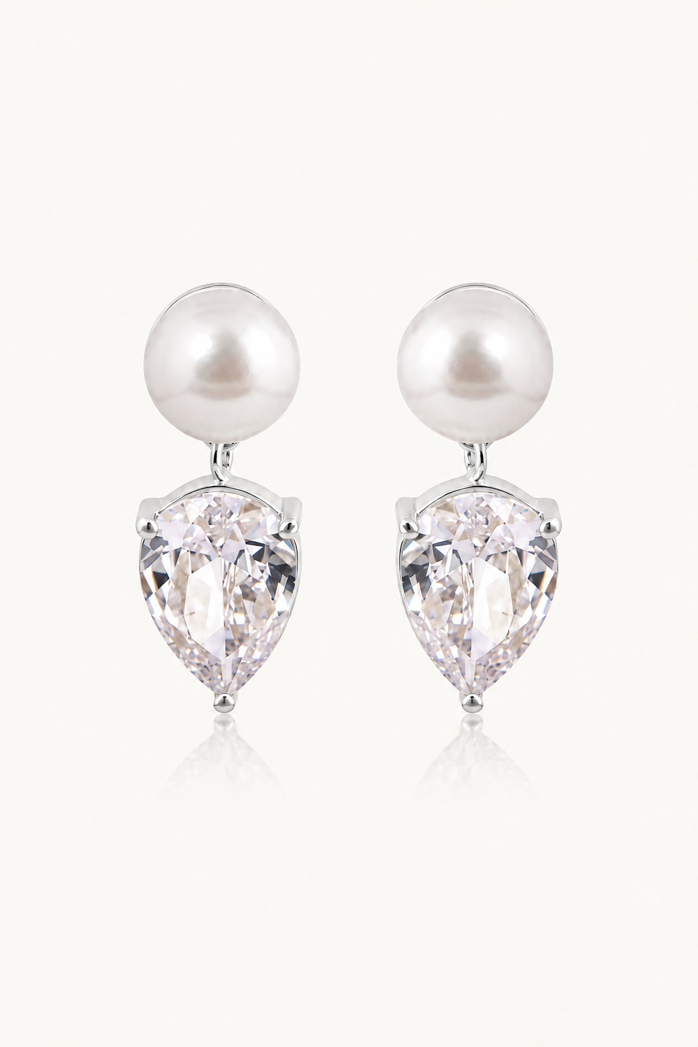 A pair of pearl bridal drop earrings featuring a white freshwater pearl and a clear pear-cut crystal in a silver-tone setting, displayed on a light cream background.