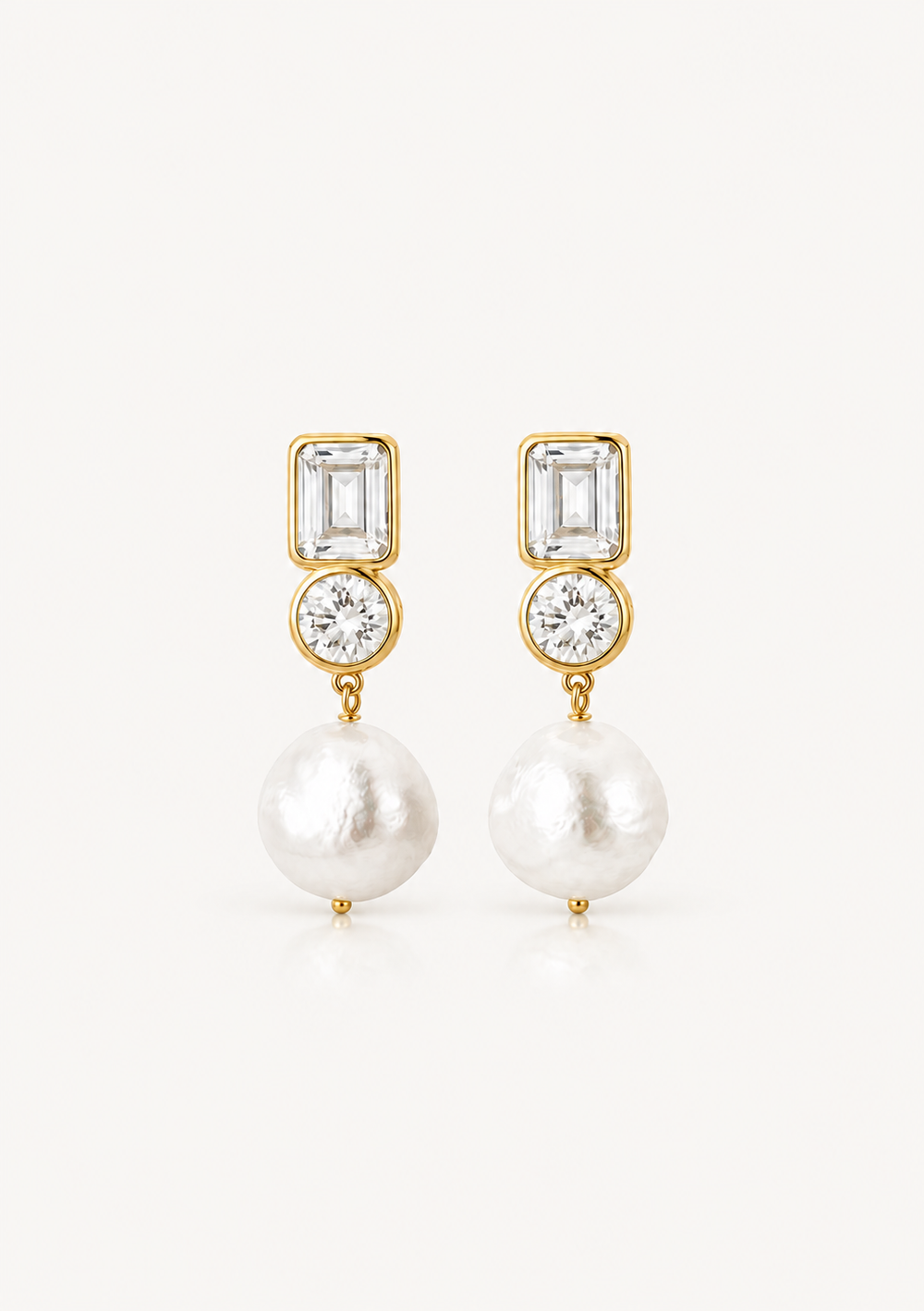A pair of gold drop earrings featuring a rectangular clear baguette-cut stone, a round clear cubic zirconia, and a large white baroque pearl dangling below, set against a plain off-white background.