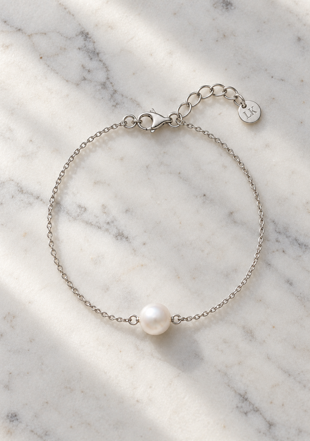 A delicate silver chain bracelet featuring a single white pearl bead, displayed on a white and gray marble surface with soft shadows.