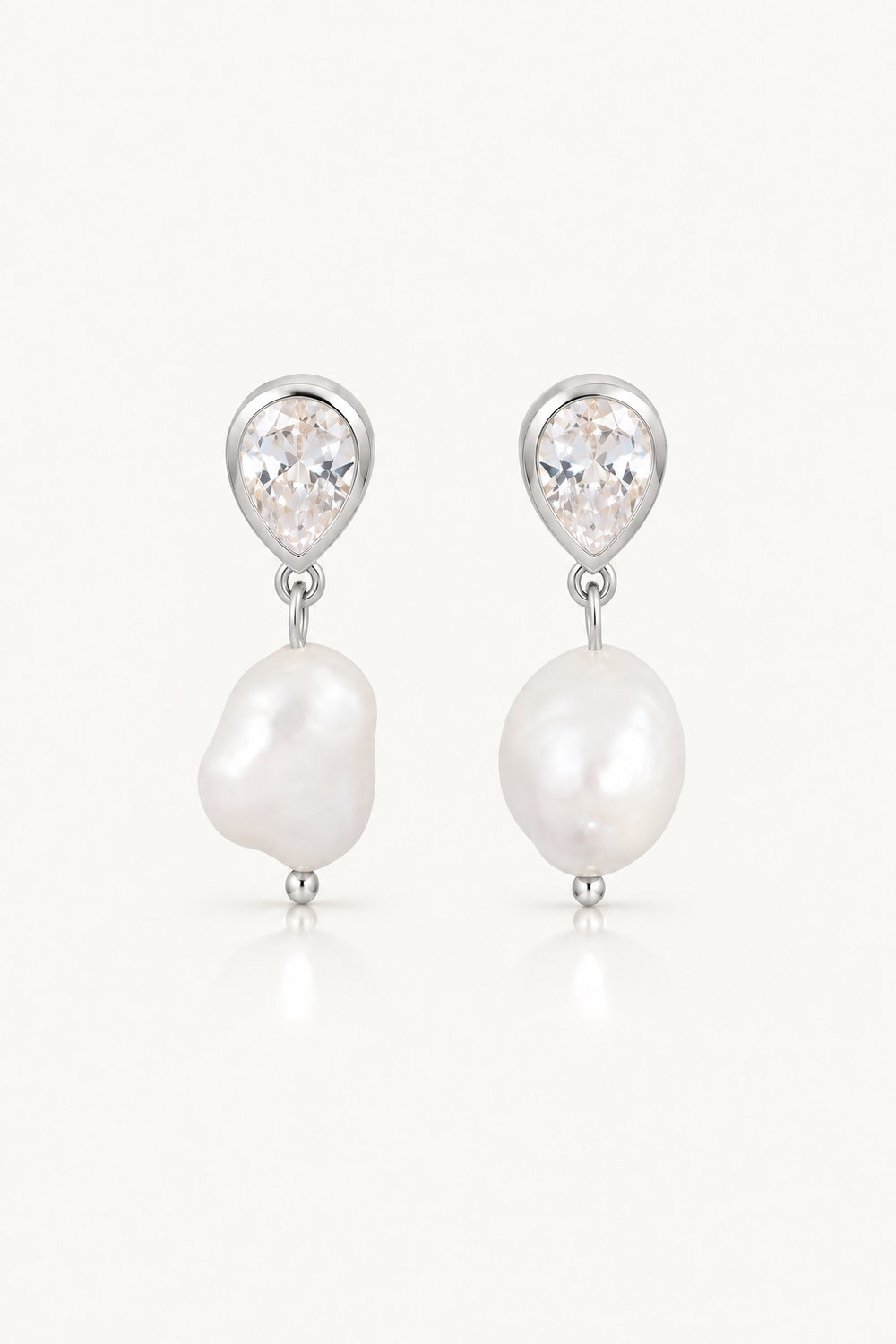 A pair of silver drop earrings featuring a small round clear crystal stud and a larger irregular white baroque pearl dangle, shown on a light cream background.