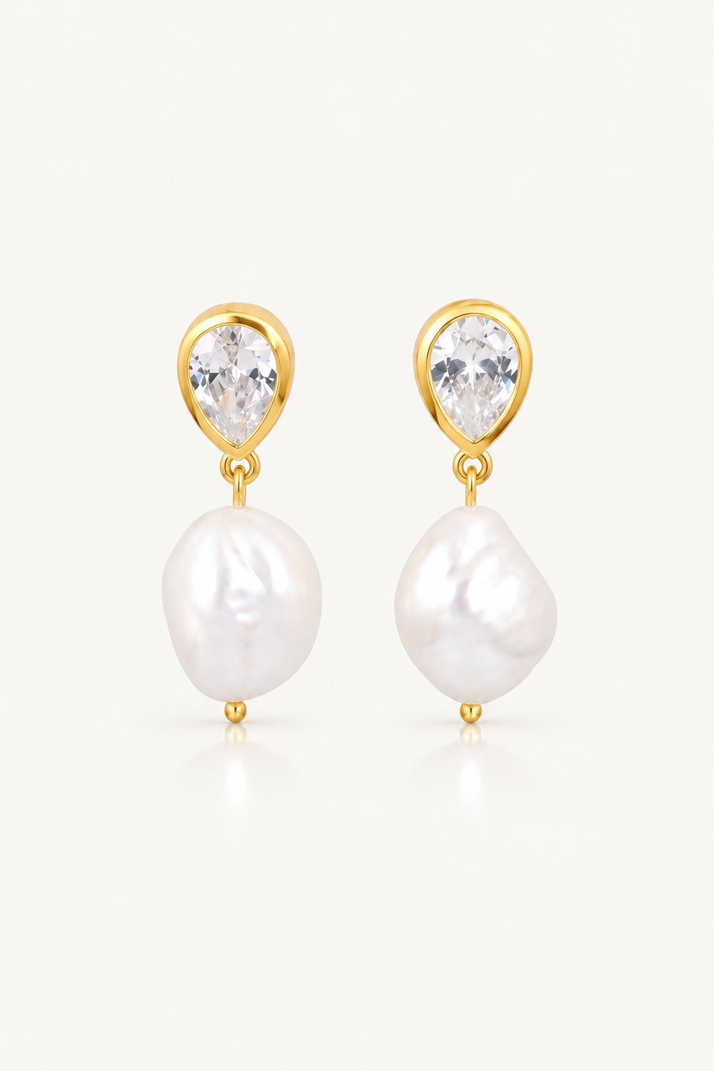 A pair of gold drop earrings featuring a small clear teardrop gemstone above a larger irregular white baroque pearl, set against a clean off-white background.