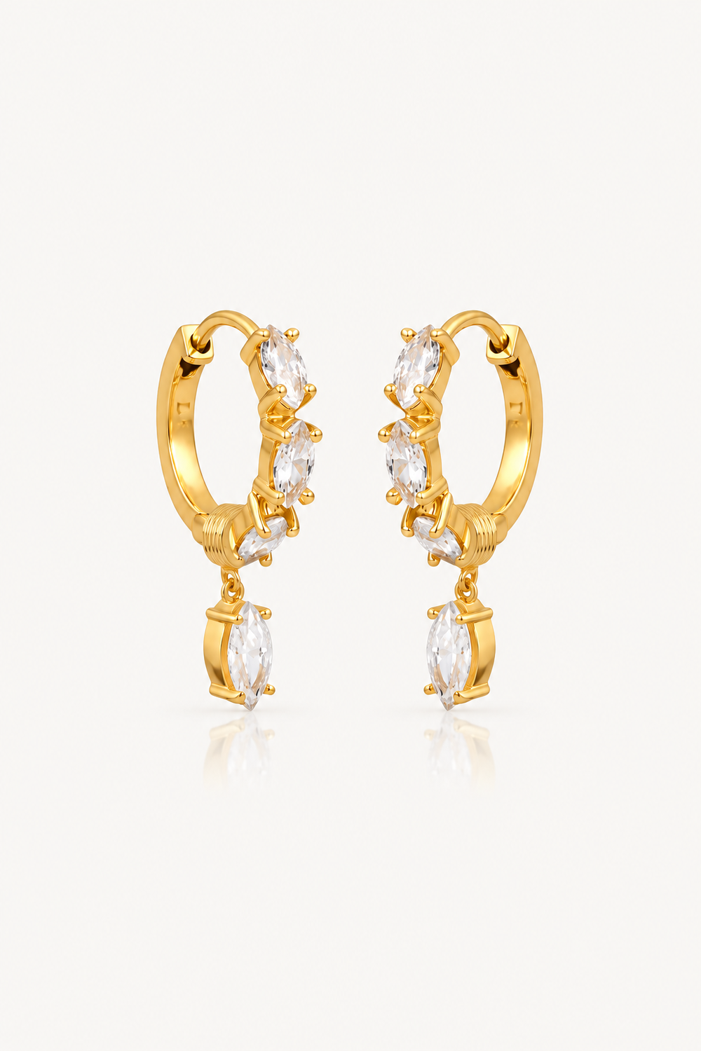 A pair of gold huggie hoop earrings with dangling clear marquise-cut crystals, shown on a light gray background with a soft reflection below.
