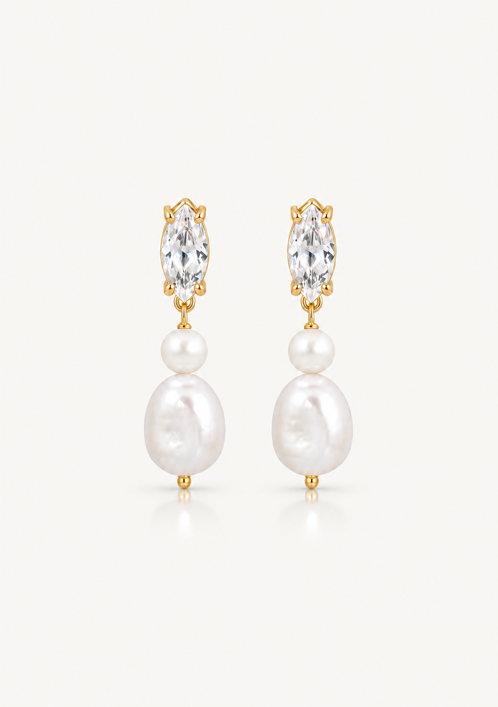 A pair of gold drop earrings featuring a marquise-cut clear crystal, a small white pearl, and a larger irregular white baroque pearl, shown on a light off-white background.