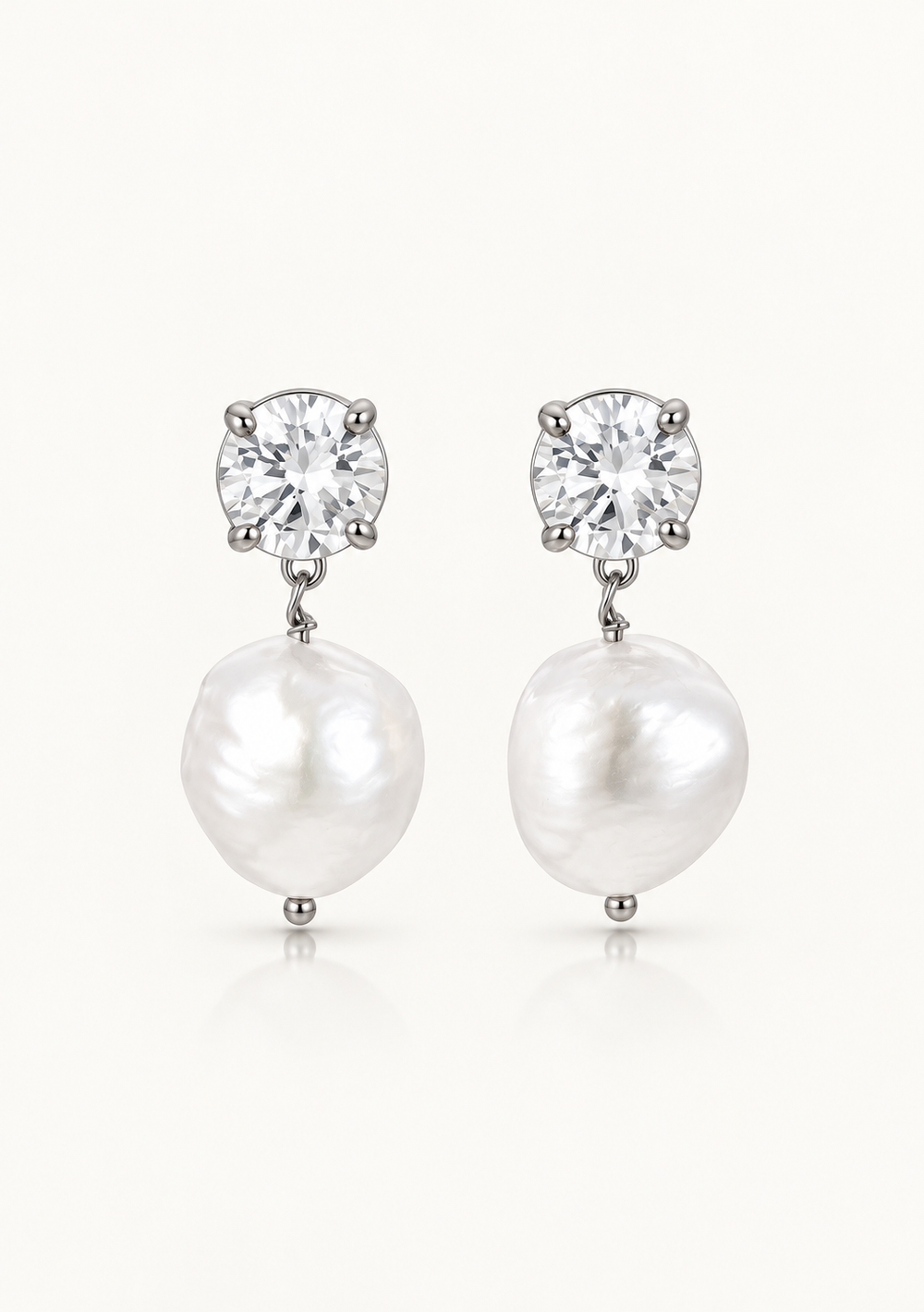 A pair of silver pearl drop wedding earrings featuring a round clear gemstone in a prong setting above a large, irregular white baroque pearl, shown on a light off-white background.