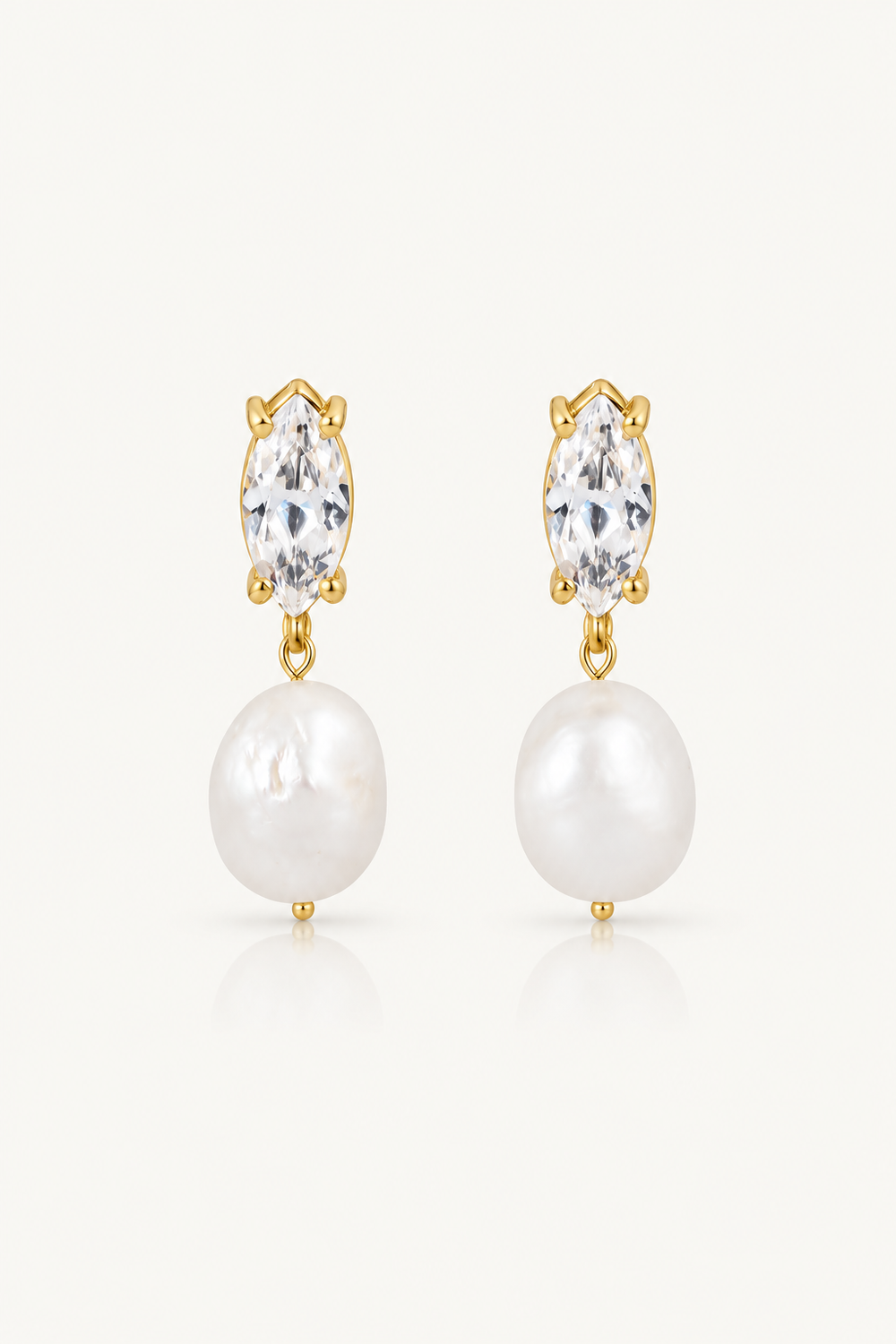 A pair of gold pearl drop bridal earrings featuring a marquise-cut clear crystal and a round white pearl, set against a clean off-white background. Made for the modern bride.