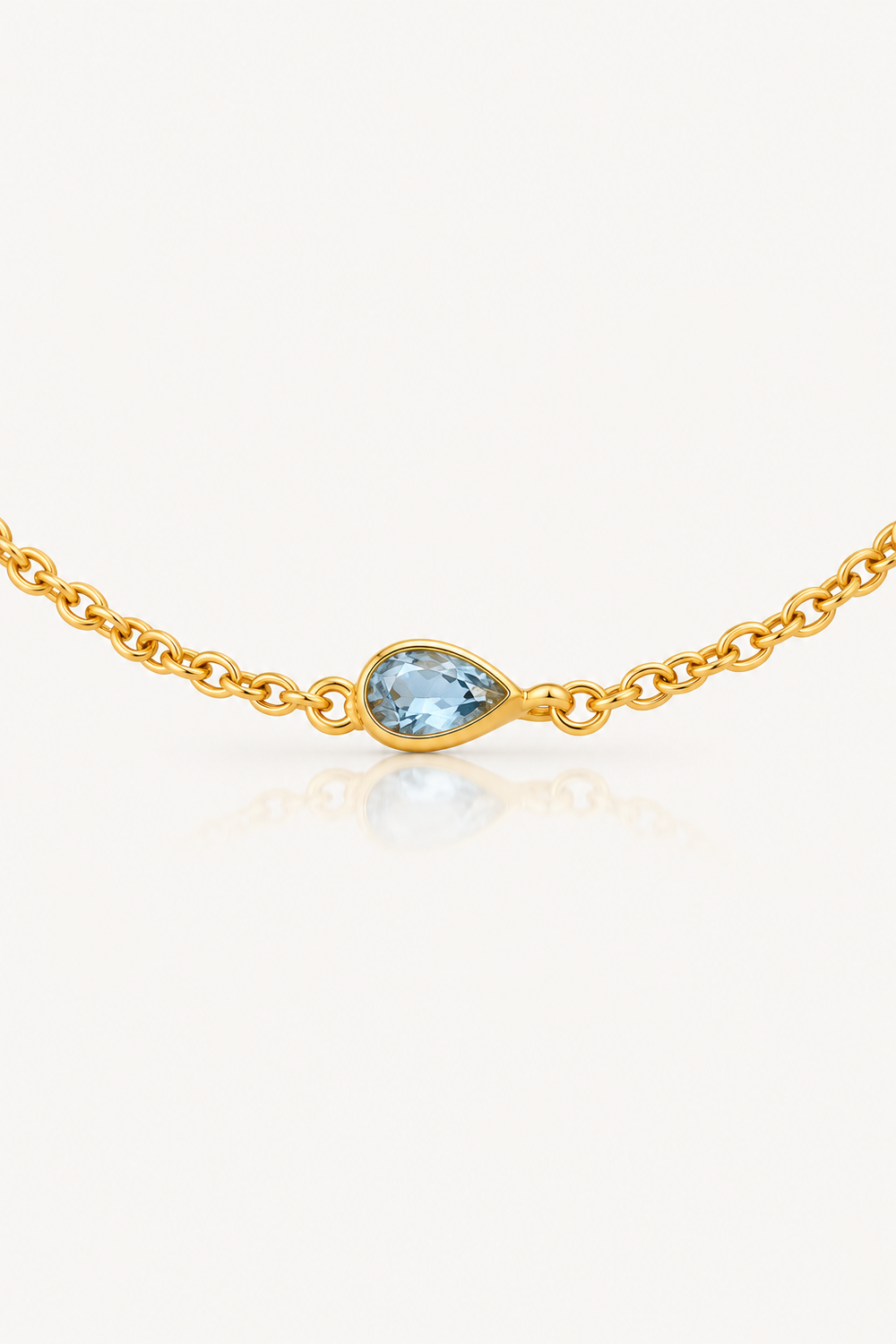 A delicate gold chain bracelet featuring a small, teardrop-shaped light blue gemstone set in a polished gold bezel, displayed on a clean off-white background with a soft reflection.