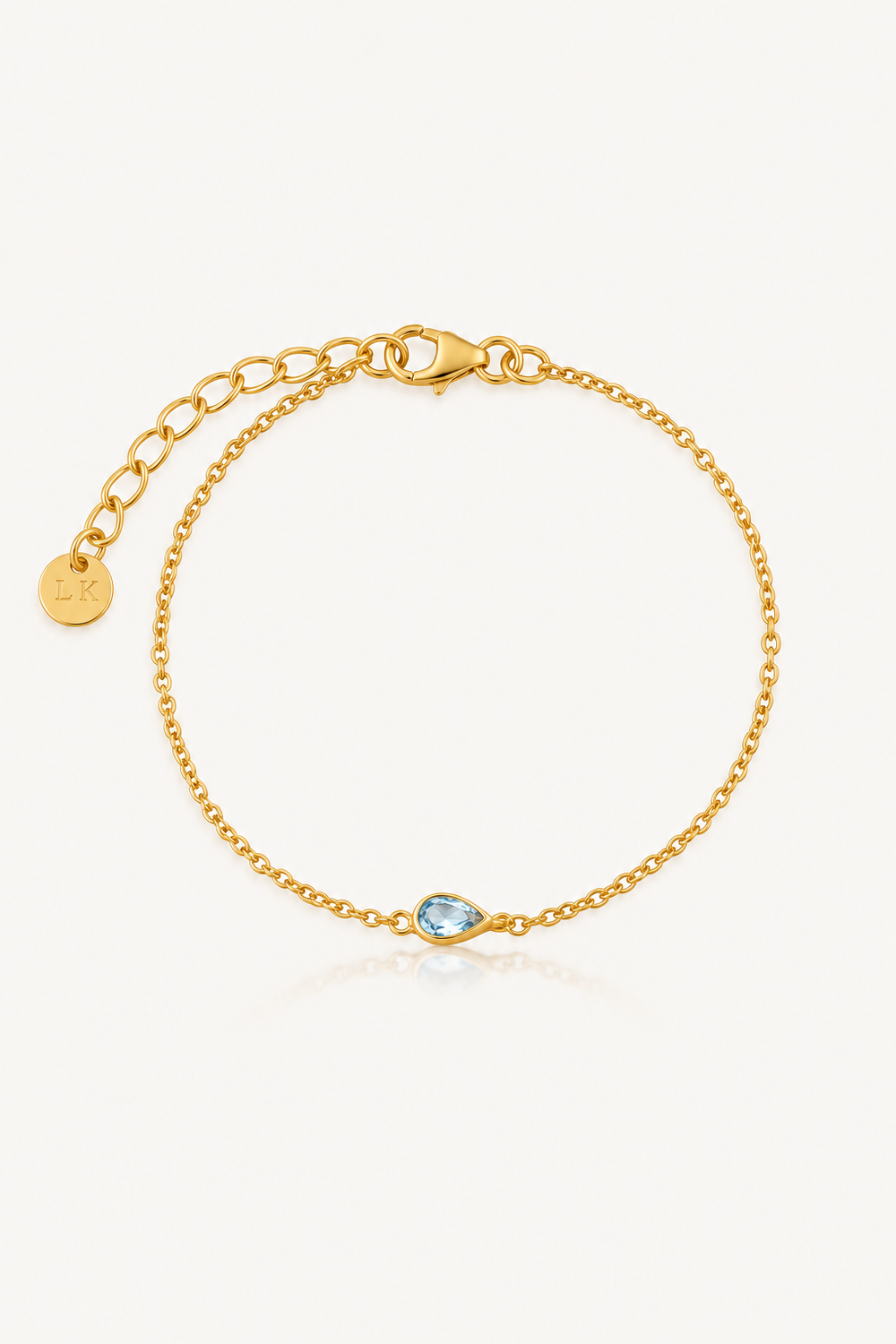 A delicate gold chain bracelet with a small teardrop-shaped light blue gemstone at the center, displayed on a clean off-white background.