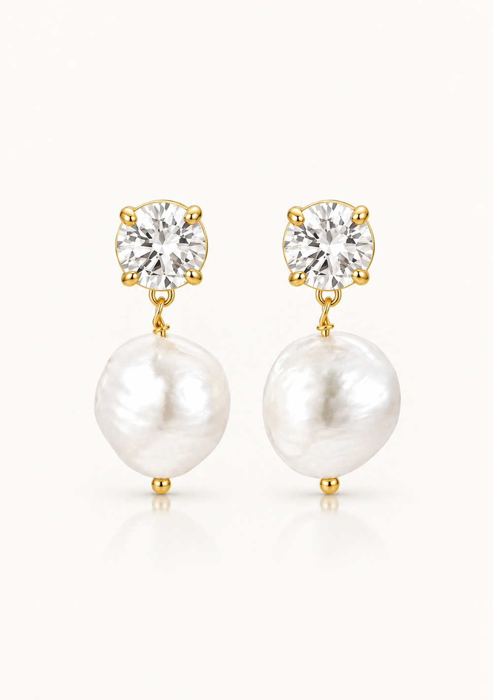 A pair of gold pearl drop wedding earrings featuring a round clear gemstone in a prong setting above a large, irregular white baroque pearl, set against a clean off-white background.