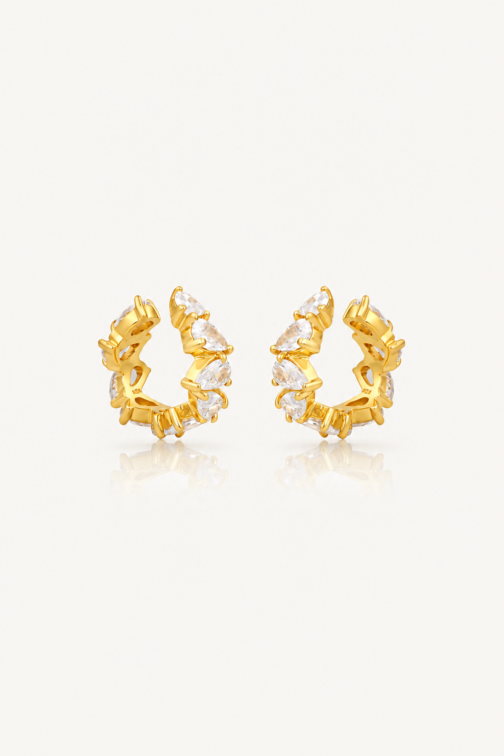 A pair of gold huggie hoop earrings adorned with clear marquise-cut gemstones, displayed on a clean off-white background with a soft reflection below.