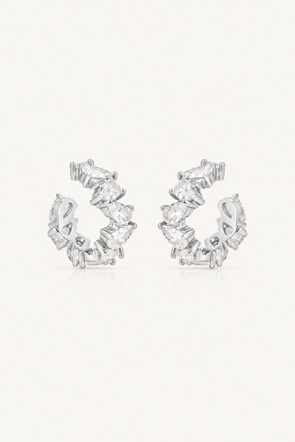 A pair of silver-toned huggie hoop earrings adorned with marquise-cut clear crystals, set against a plain off-white background.